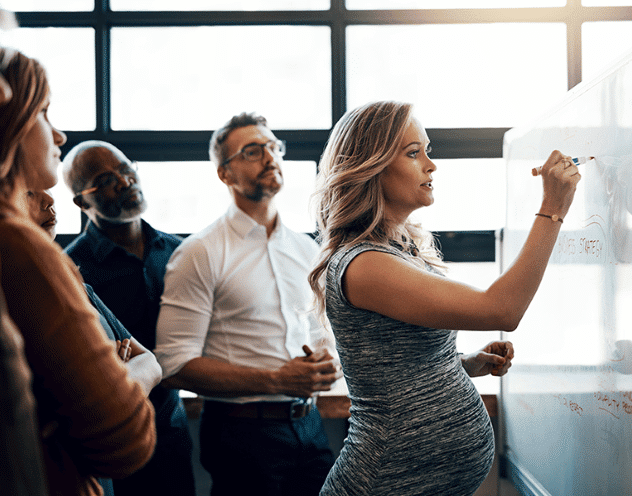 Unconscious Bias is indicated as a very pregnant woman executive leads a standing team meeting of 4 racially diverse members - 2 male and 2 females. The pregnant leader writes “Strategy” on a white board.