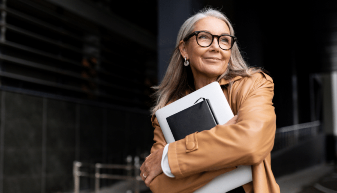 Female Business professional wearing glasses while holding laptop and notebook close to chest and looking hopeful with a smile staring in the distance walking into an office building garage.