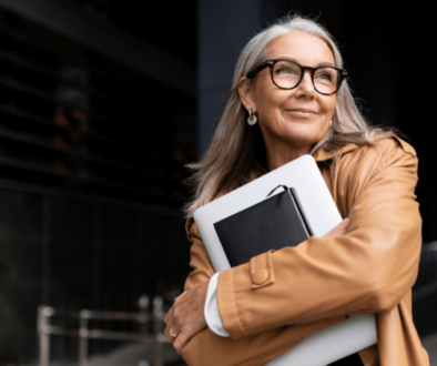 Female Business professional wearing glasses while holding laptop and notebook close to chest and looking hopeful with a smile staring in the distance walking into an office building garage.