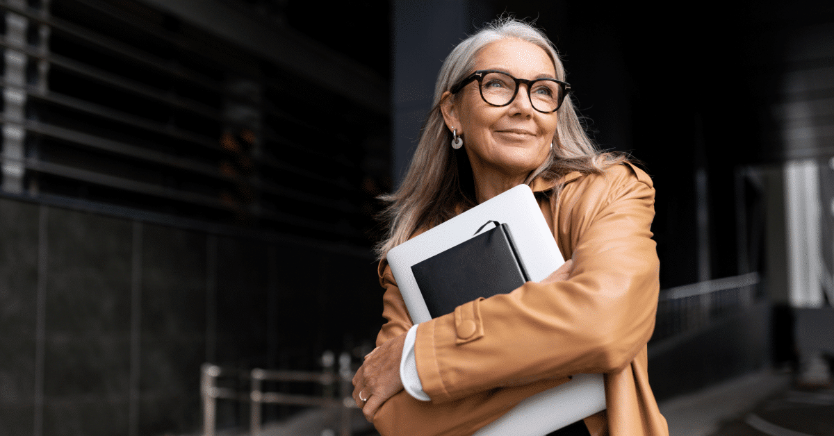 Female Business professional wearing glasses while holding laptop and notebook close to chest and looking hopeful with a smile staring in the distance walking into an office building garage.